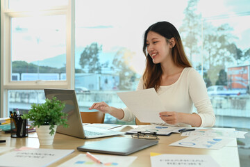 Pleasant young female employee using laptop computer and checking financial data at her office desk