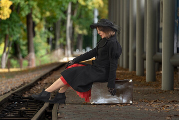 Beautiful woman in black coat and hat sits on leather suitcase waiting for train in station. Photo in style of 30-40s.