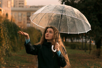 Young woman standing under transparent umbrella watches to see if it is raining. Girl with brown hair walks in fall park