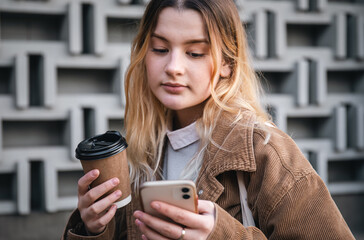 A young woman with a cup of coffee and a smartphone on the background of a wall.