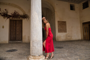 Fototapeta na wymiar Young, attractive, blonde woman in an elegant red party dress leaning on a marble column smiling happily. Concept beauty, fashion, elegance, luxury.