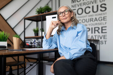 Middle aged older business woman in the office at the table looks thoughtfully into the distance