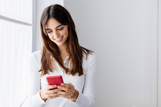 Smiling Woman Using Smart Phone By Window At Home