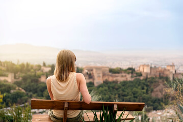 A young female traveler sitting on a bench contemplates the views of the Alhambra palace in the city of Granada, Spain