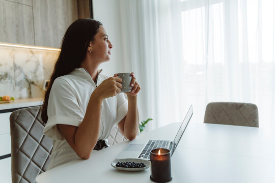 Businesswoman holding coffee cup sitting with laptop on table at home