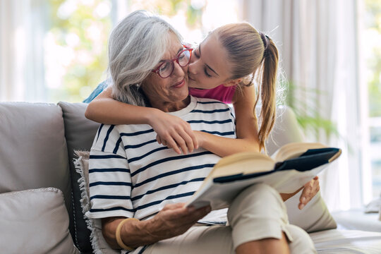Girl kissing grandmother reading book sitting on sofa at home