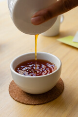 Vertical shot of tea cup being filled with hot red tea on a wooden table.