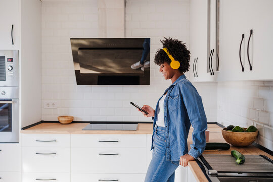 Smiling young woman wearing wireless headphones standing by kitchen counter at home