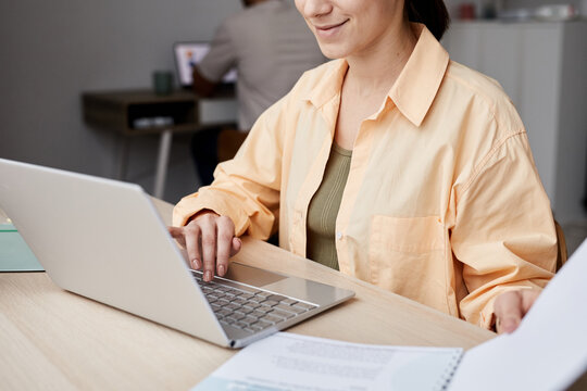 Cropped shot of young smiling female student sitting by workplace in front of laptop and preparing for project or carrying out task