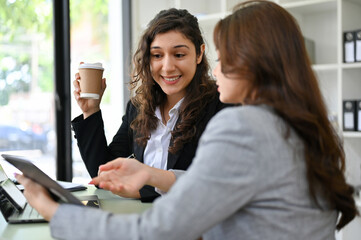 Happy millennial Caucasian businesswoman talking with her assistant while sipping coffee.