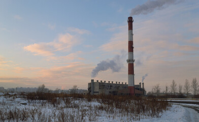 Fototapeta na wymiar Boiler room with a tall striped smoking chimney in the middle of a winter evening landscape
