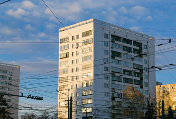 Close-up of a Soviet white high-rise building illuminated by the evening sun and many electrical wires in front of it against the blue sky