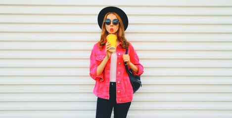 Portrait of stylish young woman drinking fresh juice wearing pink jacket, black round hat, backpack on white background