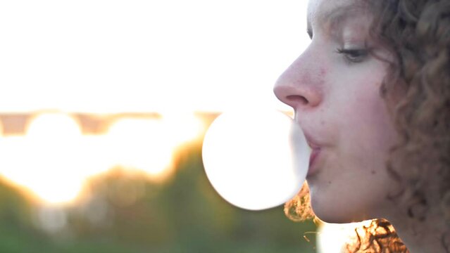 Curly-haired teenage girl inflates a big gum bubble until it bursts.