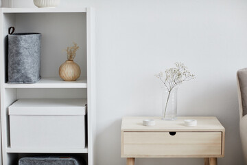 Part of cozy bedroom with dried flowers in transparent vase standing on nightstand and shelf with large white box by white wall