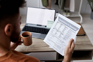 Close-up of young man with cup of tea and paper with questions in Spanish sitting by workplace and...