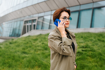 Young asian woman in stylish round glasses talking on a smartphone while walking in a modern area. Side view of a woman using a phone, calling outdoors and looking at camera
