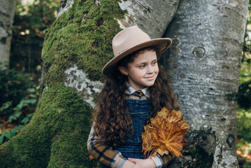 Fototapeta na wymiar Portrait of dreamful beautiful happy little child girl in hat holding maple leaves laughing and enjoying calm autumn forest during walk outdoors. Smiling kid in fall nature. People lifestyle concept