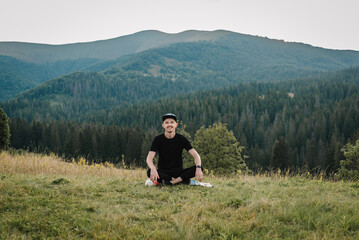 Fototapeta na wymiar Young tourist sitting on top of a mountain at sunset and enjoying a view of nature. Man relaxing on autumn grass in a field. Landscape with traveler, foggy hills, forest in fall. World Tourism Day.