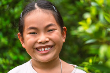 Portrait healthy Asian child girl with a big smile, standing outdoors in a park, with blurred background of the green bush, headshot of a cute little girl, space for text.