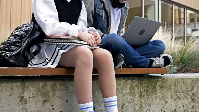 Two cute high school students, a guy and a girl are sitting on a bench in the school yard. Teenagers have fun discussing something, looking at the laptop screen.