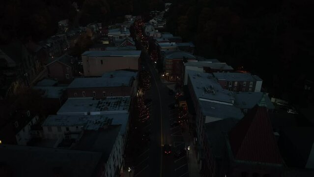Car drives through dark town at night. Descending aerial past village clock tower and Christmas tree lights illuminated for holidays.
