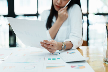 A portrait of a beautiful Asian female employee showing a stressed face while using the phone and financial documents on her desk