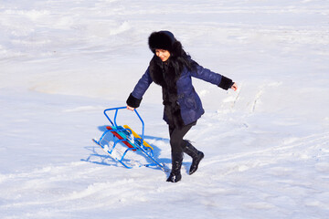 Fototapeta na wymiar Cheerful girl with a sled in a blue sheepskin coat walking in the snow