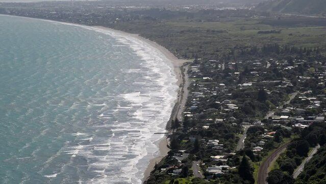 The ocean town Paekākāriki viewed from a lookout point on top of a close-by hill. Handheld tilting shot of long waves crashing ashore in midday sunlight