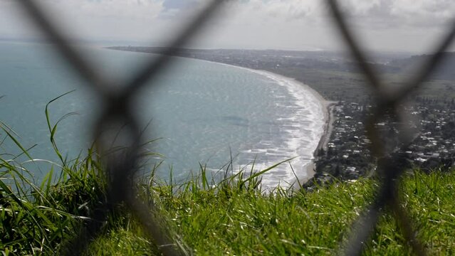 Grassy edge of a mountain towering above a oceanfront town with chain-link fence in foreground