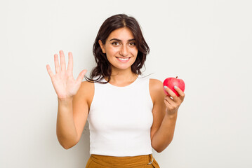 Young Indian woman holding an apple isolated on white background smiling cheerful showing number...