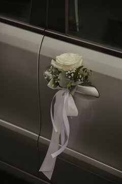 A close-up of a wedding car decorated with a white rose, greenery, and a flowing ribbon.