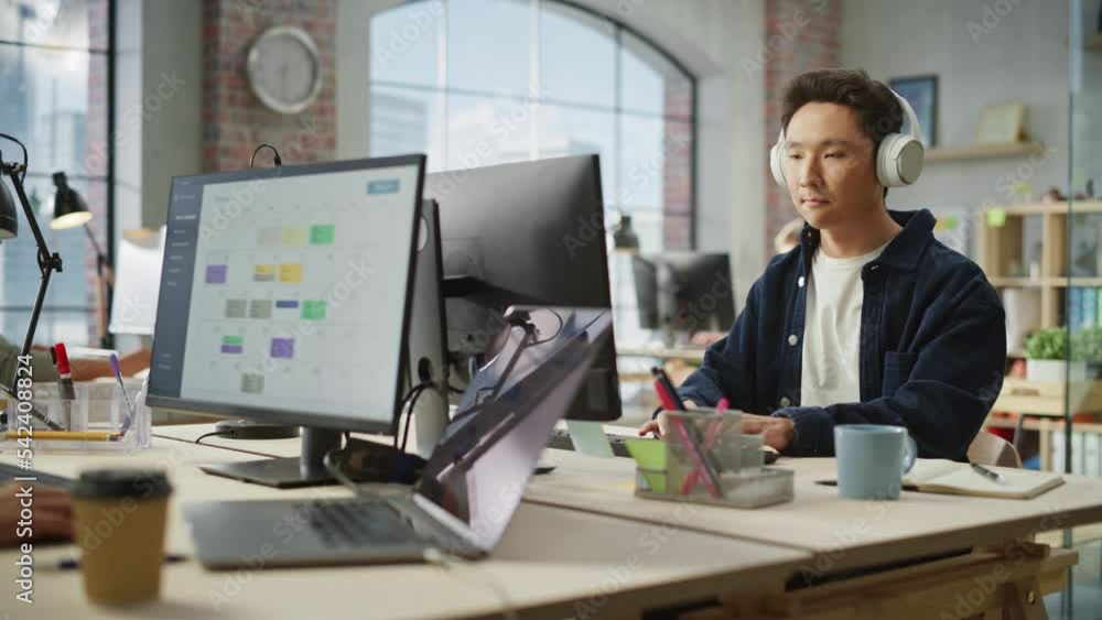 Poster portrait of asian creative focused man smiles while using computer and headphones in bright modern o