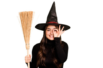 Young caucasian woman dressed as a witch holding a broom isolated on green chroma background excited keeping ok gesture on eye.