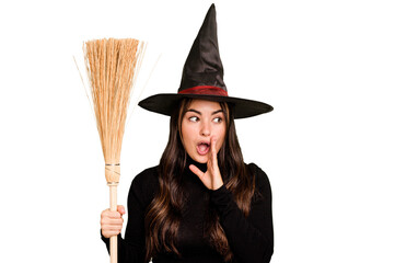 Young caucasian woman dressed as a witch holding a broom isolated on green chroma background is saying a secret hot braking news and looking aside