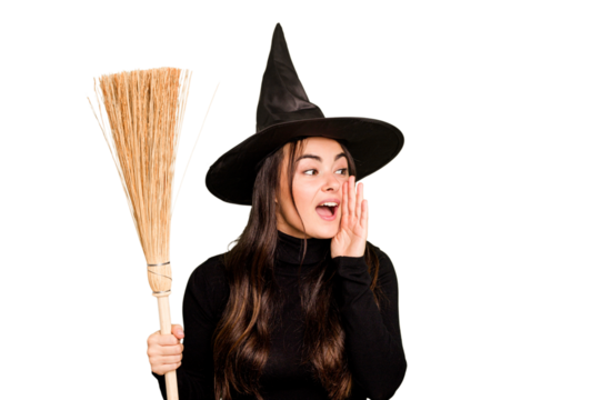 Young caucasian woman dressed as a witch holding a broom isolated on green chroma background shouting and holding palm near opened mouth.