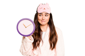 Young caucasian woman holding a clock wearing a pajama isolated on green chroma background confused, feels doubtful and unsure.