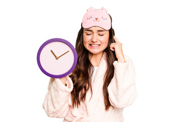 Young caucasian woman holding a clock wearing a pajama isolated on green chroma background covering ears with hands.