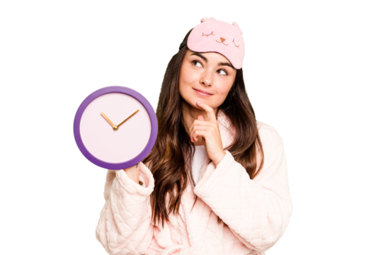 Young caucasian woman holding a clock wearing a pajama isolated on green chroma background looking sideways with doubtful and skeptical expression.