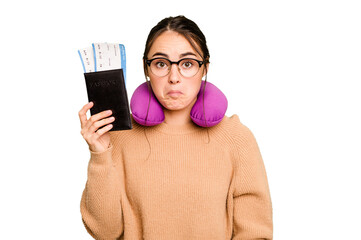 Young caucasian woman with inflatable travel pillow holding passport isolated on green chroma background shrugs shoulders and open eyes confused.