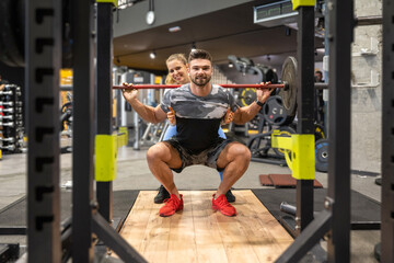 Muscular handsome man in squat position lifting heavy weights with assistance of young attractive fit woman at the gym.