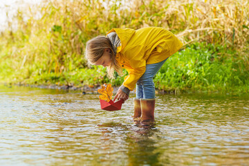 happy child girl in a yellow raincoat and paper boat at the river in autumn on nature.