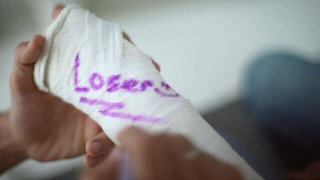 Close-up high-angle view of unrecognizable man writing joking inscription on cast of friend with broken hand wrapped in plaster bandage. Concept of friendship and fun. Shooting in slow motion.