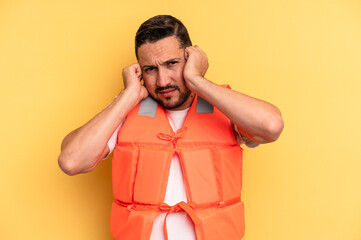 Young latin beach watcher wearing a lifejacket isolated covering ears with hands.