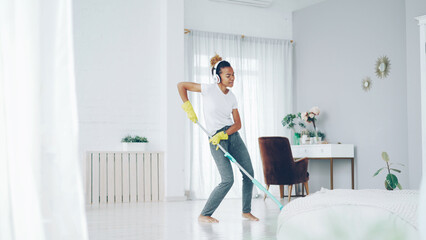 Fototapeta na wymiar Cheerful African American girl in gloves is mopping floor, listening to music with wireless headphones and dancing enjoying rhythm cleaning her nice modern apartment.