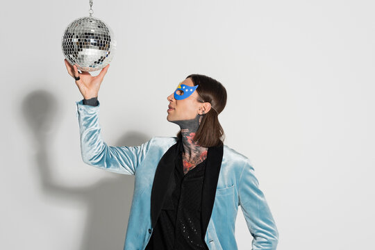 Queer Person In Elegant Silk Blazer And Party Mask Touching Shiny Disco Ball On Grey Background