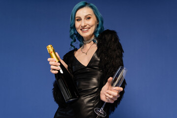 trendy woman in black faux fur jacket and leather dress holding champagne and smiling at camera isolated on blue