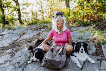 Naklejka na ściany i meble Senior woman having break during walking her three dogs in forest.