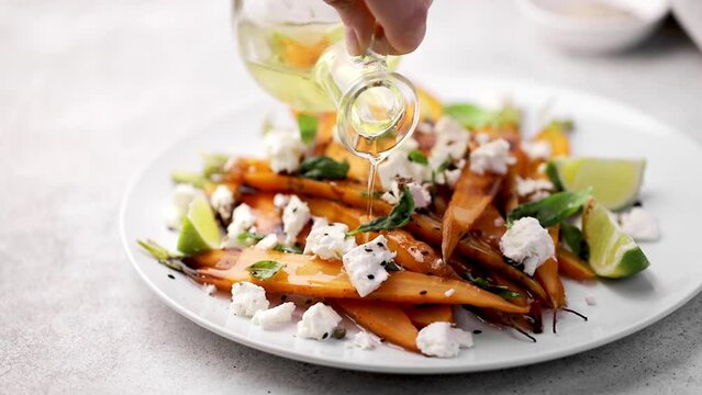Pouring oil into a salad with roasted baked carrots and feta cheese, herbs on white plate, grey background. Vegetable salad with roasted young carrots, feta cheese.