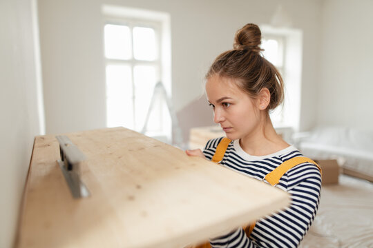 Young woman remaking her apartment, measuring wall with spirit level and hanging shelf.